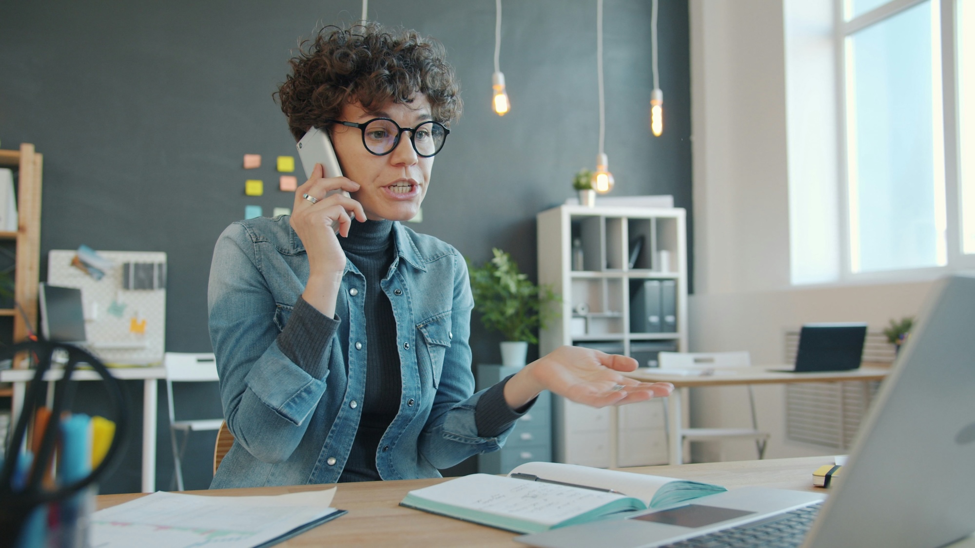 Unhappy businesswoman is yelling on phone call in office talking on mobile sitting at desk alone. 
